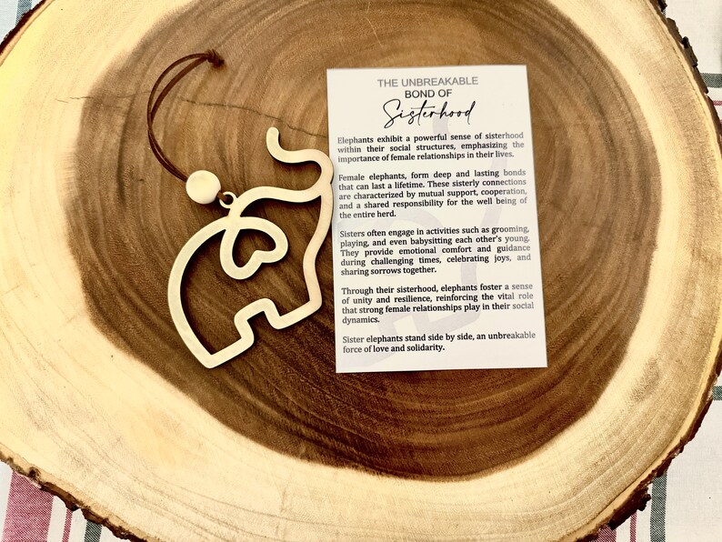May include: A wooden elephant ornament with a heart-shaped cutout hangs from a brown cord. The ornament is on a wooden surface. The text on the card reads: "The Unbreakable Bond of Sisterhood. Elephants exhibit a powerful sense of sisterhood within their social structures, emphasizing the importance of female relationships in their lives."