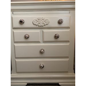 May include: White dresser with five drawers and decorative floral accents. Each drawer has a round knob with a floral pattern.