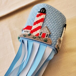 May include: A blue and white canvas craft project with a lighthouse design and blue and white ribbons attached.