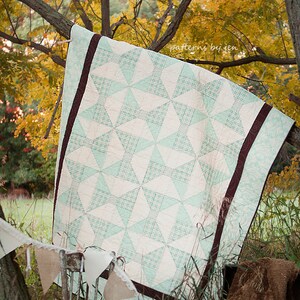 Pode incluir: Um edredom branco e verde com uma borda marrom pendurado em um galho de árvore em um cenário de outono. O edredom tem um padrão de moinho de vento e o texto "patterns by jen" é visível no canto superior direito.