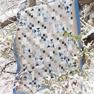 May include: A blue and beige patchwork quilt with a geometric pattern, hanging on a snowy branch in a wintery forest.