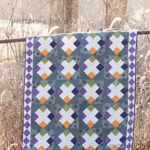 May include: A quilt with a geometric pattern of squares and diamonds in shades of purple, green, orange, and white. The quilt is hanging on a fence.