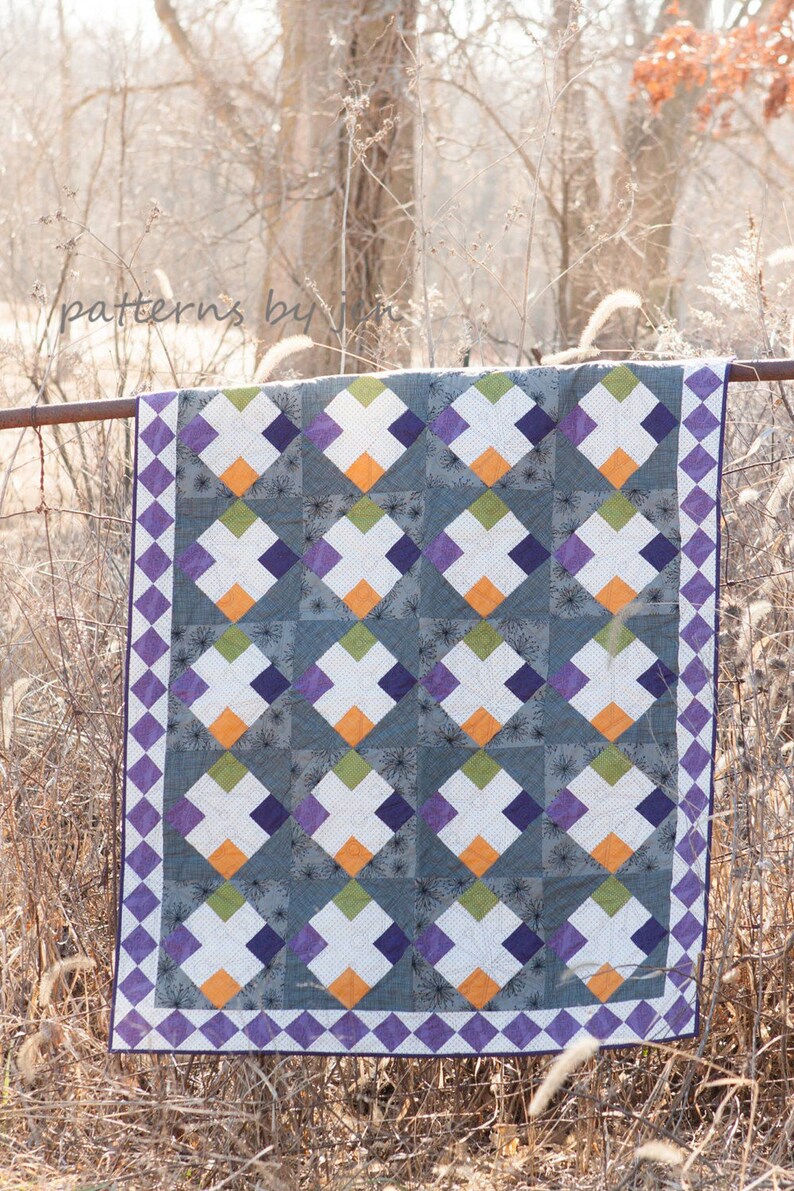 May include: A quilt with a geometric pattern of squares and diamonds in shades of purple, green, orange, and white. The quilt is hanging on a fence.