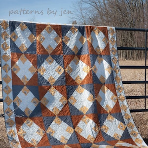 May include: A quilt with a geometric pattern in shades of blue, orange, and yellow. The quilt is hanging on a black metal fence against a backdrop of a blue sky and bare trees. The text "patterns by jen" is visible at the top of the image.