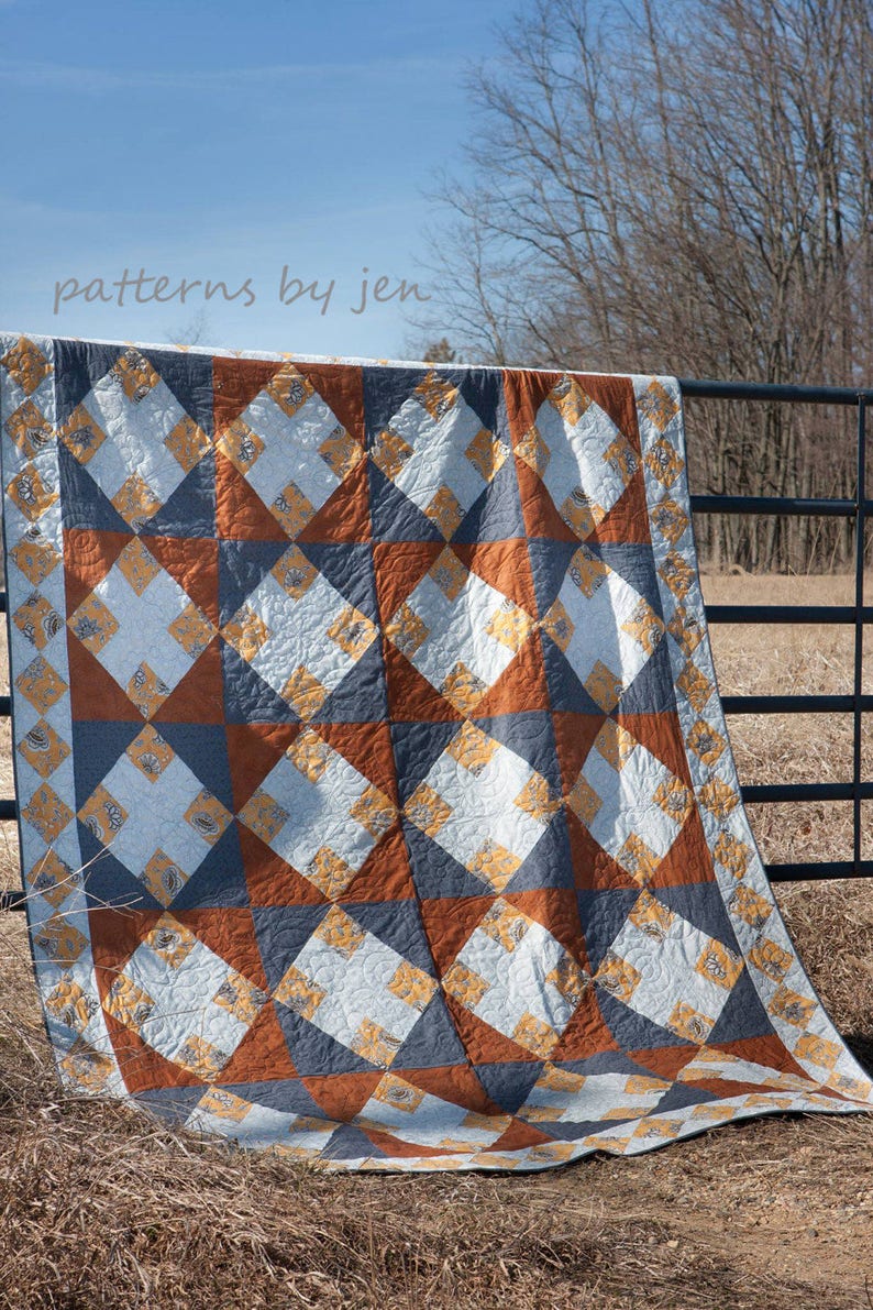 May include: A quilt with a geometric pattern in shades of blue, orange, and yellow. The quilt is hanging on a black metal fence against a backdrop of a blue sky and bare trees. The text "patterns by jen" is visible at the top of the image.
