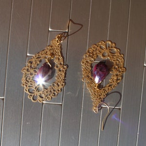 May include: A pair of gold-colored filigree earrings with a teardrop-shaped purple gemstone. The earrings have a delicate, openwork design and are displayed on a silver, textured surface. The gemstones catch the light, creating a sparkling effect.
