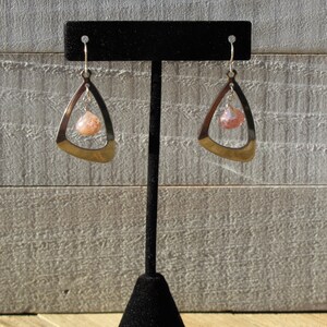 May include: A pair of silver earrings with a pink and white stone hanging from a triangle-shaped frame.