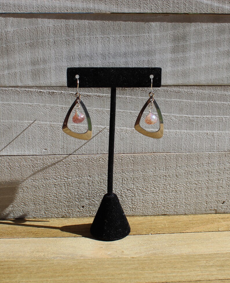 May include: A pair of silver earrings with a teardrop-shaped design and a pink gemstone dangle. The earrings are displayed on a black earring stand.