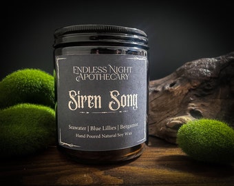 May include: A dark glass jar candle with a black lid and a label that reads "Endless Night Apothecary Siren Song." The label lists the scent notes: Seawater, Blue Lillies, and Bergamot. The candle is surrounded by green moss.