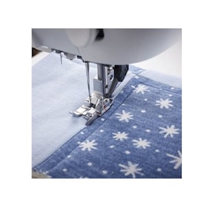 May include: A close-up of a sewing machine in operation, with the needle and presser foot visible. The machine is stitching two pieces of fabric together. The fabric on the right is blue with white snowflake patterns, and the fabric on the left is light blue.