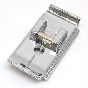May include: A shiny, silver-colored sewing machine foot with a rectangular shape. The foot has a small, cylindrical gold-colored pin and the numbers "412 6870" are visible. The foot is designed to attach to a sewing machine.