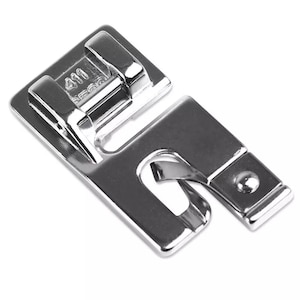 May include: A shiny, silver sewing machine foot with the number "411" stamped on the top. The foot has a rectangular shape with a curved opening and a small, square end. It is designed to attach to a sewing machine.