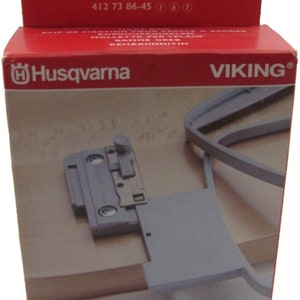 Può includere: Una scatola rossa Husqvarna Viking con un accessorio per macchina da cucire. La scatola presenta le scritte "Husqvarna" e "Viking". L'accessorio è grigio e sembra essere realizzato in metallo e plastica. La scatola riporta anche la scritta "KEEPING THE WORLD SEWING".
