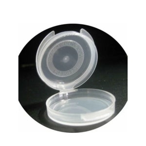 May include: A clear, hinged plastic container, open to reveal the interior. The lid is attached to the base by a hinge. The container is circular and appears to be made of a transparent material. The text on the lid is blurry.