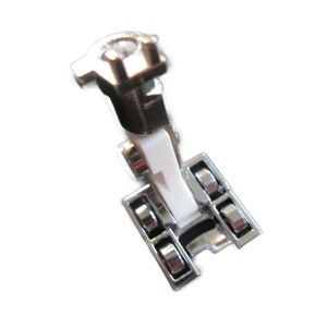 May include: A close-up of a sewing machine foot, made of shiny silver metal and white plastic. The foot has a rectangular base with multiple rollers and a top section with a mounting hole. The item is designed for sewing.