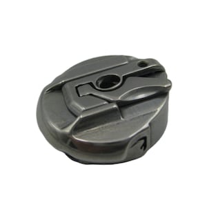 May include: A close-up of a metallic grey sewing machine bobbin case. The case has a circular shape with a complex design featuring various grooves and openings. The metal has a slightly textured appearance, and the overall aesthetic is industrial.