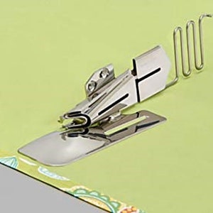 May include: A shiny, silver-colored sewing machine binder attachment. The attachment is on a light green surface with a patterned fabric edge. The binder has a curved metal guide and a flat base, designed to fold fabric edges.