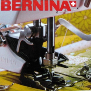 May include: Close-up of a Bernina sewing machine with the brand name in red. The machine's needle and presser foot are visible, along with other metal components. The background is a yellow and green floral pattern.