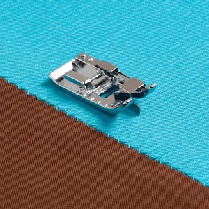 Puede incluir: Un prensatelas brillante de máquina de coser plateado. El prensatelas está colocado sobre una costura diagonal de tela marrón y turquesa. La tela tiene un borde cosido. El prensatelas está diseñado para sujetar la tela mientras se cose.