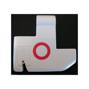 May include: A white, T-shaped object with a red circle and a rectangular inset. The object has a small, curved extension on one side. The background is black.