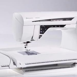 Pode incluir: Máquina de costura Husqvarna Viking branca com um grande ecrã tátil. A máquina tem uma mesa de extensão integrada e uma variedade de botões e controlos. O design inclui detalhes florais decorativos na lateral.