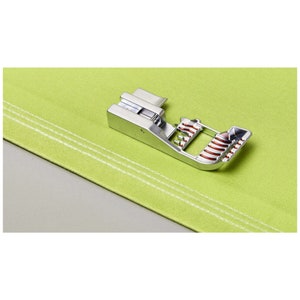 Puede incluir: Un prensatelas plateado para máquina de coser con detalles rojos, colocado sobre una tela verde claro con costuras blancas. El prensatelas está diseñado para coser y es de metal.