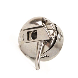 May include: A close-up of a silver-colored sewing machine bobbin case. The case has a curved shape with a rectangular opening and a curved metal lever. The bobbin case is designed to hold the bobbin and thread for sewing machines.