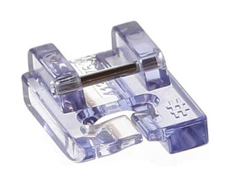 May include: A clear, plastic sewing machine foot with a metal bar. The foot has a rectangular shape with a curved opening at the bottom and a series of small, square-shaped markings on the side. It is designed to attach to a sewing machine.