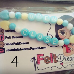 Könnte beinhalten: Ein blaues und weißes Perlenarmband mit den Wörtern "OPEN" und "CLOSE" darauf. Das Armband ist handgefertigt und hat einen Herzanhänger. Das Bild zeigt auch eine Visitenkarte mit dem Namen "Felt Dreams" und Kontaktinformationen.