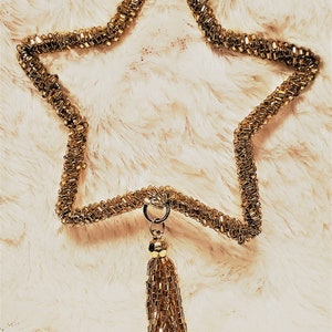 May include: A gold beaded star ornament with a tassel. The star is made of small, rectangular beads and hangs from a gold ribbon. The tassel is made of similar beads. The ornament is displayed against a fluffy, white background.