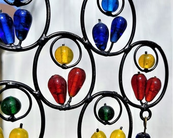 May include: A colorful glass bead wind chime with a black metal frame. The beads are arranged in a circular pattern and are suspended from the frame by thin wires. The beads are red, yellow, blue, and green.