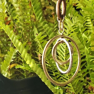 May include: Gold and silver double circle pendant necklace with a delicate chain. The pendant is hanging from a green leafy plant.