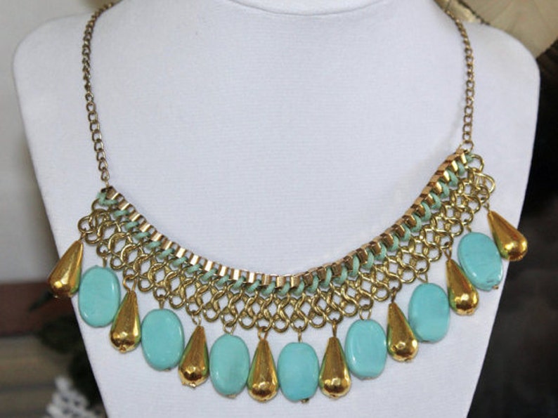 May include: A gold chain necklace with a turquoise and gold pendant. The pendant is made of multiple turquoise beads and gold chain links. The necklace is a statement piece and would be perfect for a night out.