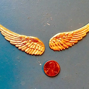May include: Two gold-coloured metal angel wings are displayed on a blue surface, with a penny placed below them for size comparison. The wings have detailed feather textures and a metallic sheen, suggesting a decorative or craft item.