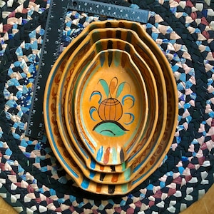 Puede incluir: Juego de cuatro cuencos de cerámica ovalados anidados con diseños pintados a mano en naranja, marrón, azul y verde. Los cuencos están colocados sobre una alfombra trenzada. Una regla está colocada junto a los cuencos.