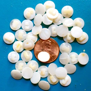 Puede incluir: Un conjunto de pequeños botones redondos blancos con brillo nacarado, dispersos sobre una superficie azul. Se incluye una moneda de cobre como referencia de tamaño. Los botones tienen un diseño claro y en relieve en el centro.