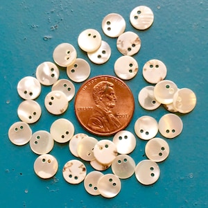 May include: A collection of small, round, off-white mother-of-pearl buttons with two holes each, scattered on a teal surface. A copper penny is included for size comparison.
