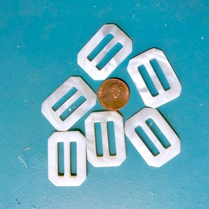 May include: Six white rectangular belt buckles with a penny in the center. The buckles have two parallel slots and a pearlescent sheen. The buckles are arranged in a circular pattern on a teal surface.