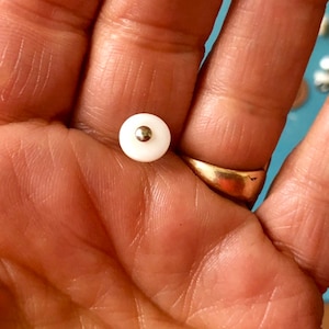 May include: A small, round, white button with a silver-colored center. The button is held in a hand, with a gold ring visible on a finger. The background is a blurred blue.