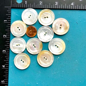 May include: A collection of ten iridescent, round buttons with two holes each, arranged on a teal surface. A penny is included for size comparison. The buttons have a pearlescent sheen with a subtle yellow-gold edge.