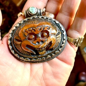 May include: An oval pendant featuring a carved brown stone face with prominent eyes and teeth, set in a textured silver frame. The pendant is topped with a small, round, light blue turquoise stone and silver beads, creating a detailed piece.