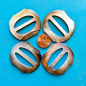 May include: Four iridescent, oval-shaped vintage buckles with a penny for scale, arranged on a bright blue surface. The buckles have a decorative, layered design and a central opening. The penny is a copper color.