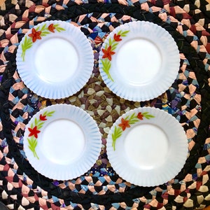 Puede incluir: Cuatro platos blancos con borde festoneado y diseño floral. Cada plato presenta una flor roja con hojas verdes. Los platos están colocados sobre una colorida alfombra trenzada.