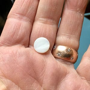 以下が含まれることがあります： 真珠のような光沢のある、小さくて丸い白いボタン。ボタンは手で持たれており、星型の装飾が施されたローズゴールドの指輪の隣にあります。