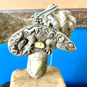Puede incluir: Un colgante de metal plateado en una cadena de bolas, con un diseño en relieve de lagarto o gecko con escamas texturizadas. El colgante incluye una pequeña incrustación de piedra amarilla ovalada y una diminuta piedra roja oscura.