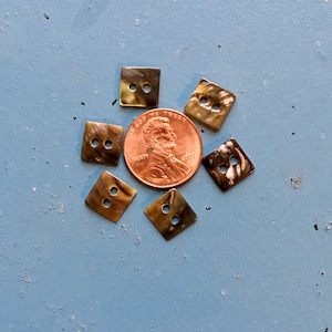 May include: Five small square buttons with a marbled brown and white pattern, each featuring two holes in the center. The buttons are arranged in a circular pattern around a single copper-colored United States one-cent coin for scale.