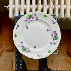 Puede incluir: Plato de porcelana blanco con borde festoneado y borde dorado. El plato está decorado con racimos de violetas moradas y hojas verdes. Una regla está al lado del plato.