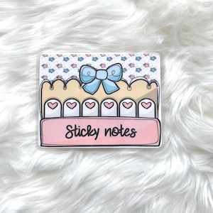 May include: A pink and blue sticky note pad with a bow and hearts. The pad says "Sticky notes" on the front.