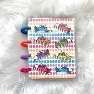 May include: A small, clear, plastic-covered notebook with a rainbow polka dot pattern and colorful cartoon sneakers. The notebook has a silver chain with a charm of a sneaker attached to it.