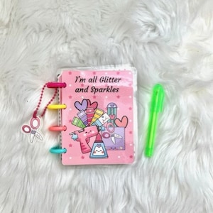 Op de afbeelding: Een roze notitieboek met een kleurrijk ontwerp met een schaar, lijm en verf. Het notitieboek heeft een transparante plastic hoes en een ringband. De tekst op de omslag luidt "I'm all glitter and sparkles".